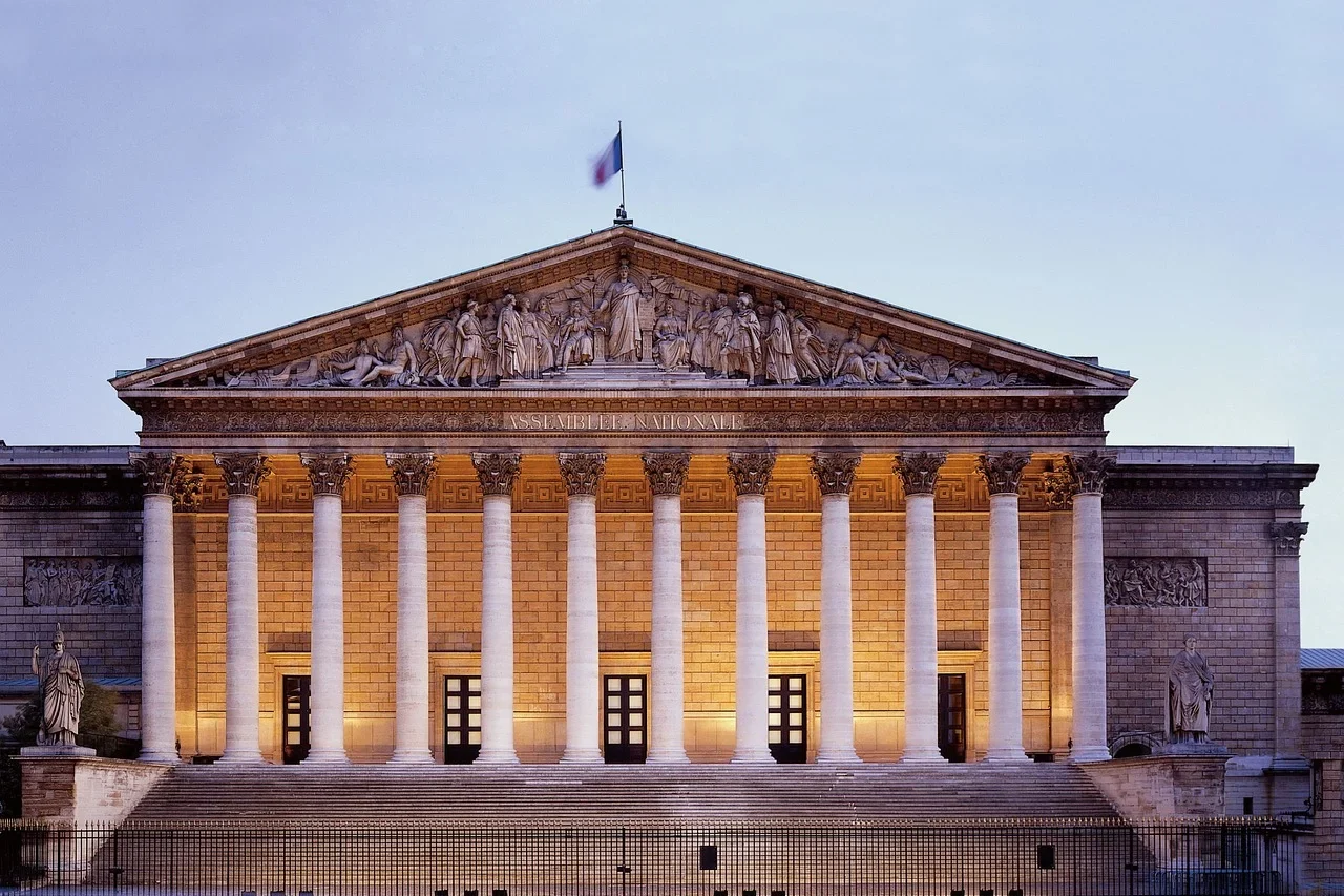 A grand neoclassical building with tall columns and intricate carvings, illuminated at dusk, with a flag atop and light trails from passing cars in the foreground which shows ideology in governance.